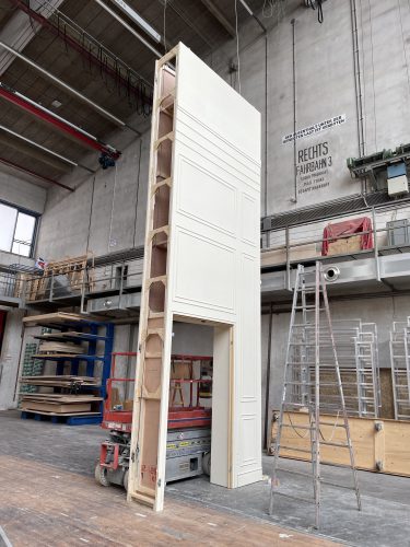 Große, weiße Kulissenwand mit Türöffnung steht in einer Werkhalle neben Leiter und Arbeitsbühne.