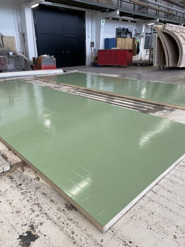 Grüne Flächen mit Kachelstruktur liegen in einer Werkhalle auf dem Boden, umgeben von Arbeitsmaterialien und Maschinen.