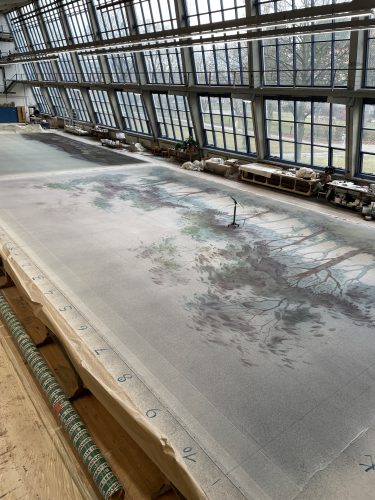 Große bemalte Leinwand mit Landschaftsmotiv liegt in einer lichtdurchfluteten Werkhalle auf dem Boden ausgebreitet.
