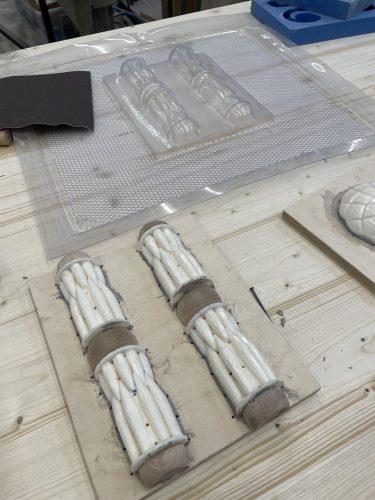 Zwei weiße Thermoformobjekte auf einer Holzplatte und eine transparente Gussform auf einem Tisch in einer Werkstatt.