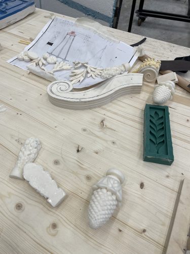 Verschiedene weiße und grüne Stuckornamente sowie Konstruktionszeichnungen liegen auf einem Holztisch.