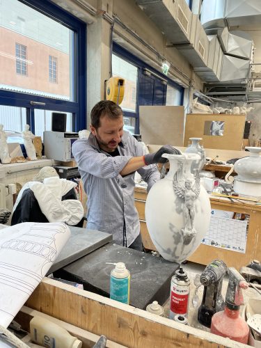 Ein Mann arbeitet in einer Werkstatt an einer verzierten Vase aus weißem Material, umgeben von Werkzeugen und Zeichnungen.