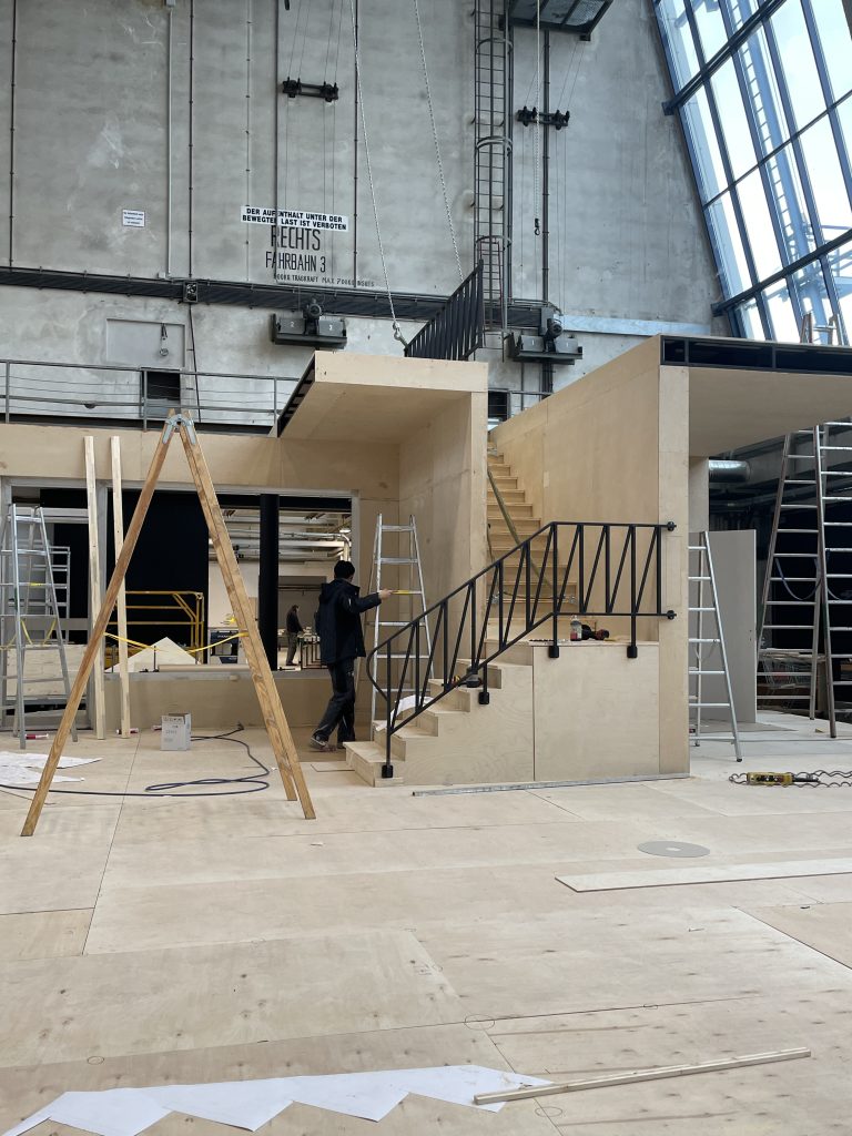 Eine Person arbeitet an einem großen Holzaufbau mit Treppe und Geländer in einer Industriehalle, umgeben von Leitern und Baumaterial.