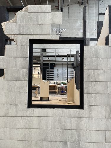 Ein rechteckiges Fenster in einer unvollständigen Betonwand ermöglicht den Blick auf eine große Werkstatt mit verschiedenen Materialien.