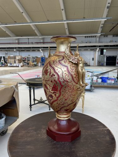 Goldene und rote Vase mit filigranem Muster und einer goldenen Figur am Rand, auf einem Holztisch in einer Halle.