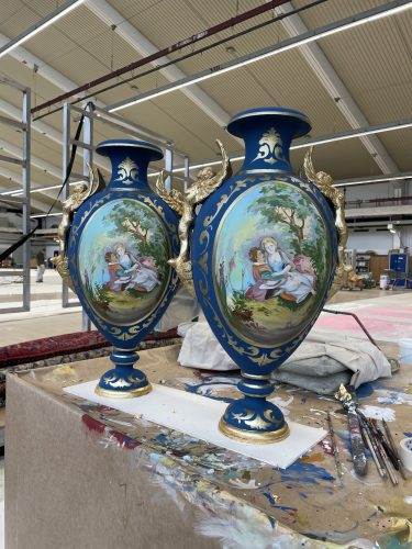 Zwei blau-goldene Vasen mit gemalten romantischen Szenen und goldenen Details stehen auf einem Arbeitstisch in einer Werkstatt.