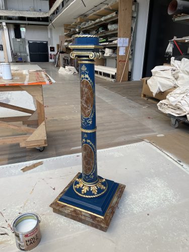Eine blau-goldene dekorative Säule mit marmorierten Verzierungen steht in einer Werkstatt neben einer Dose Holzlack.
