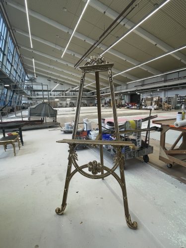 Vergoldete Staffelei mit dekorativen Ornamenten steht in einer großen Werkhalle mit Tischen und Werkstätten im Hintergrund.