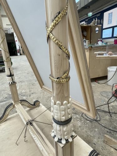 Ein kunstvoll geschnitzter Holzpfosten mit goldfarbenem, verziertem Band, Werkzeuge und Werkstatt im Hintergrund.