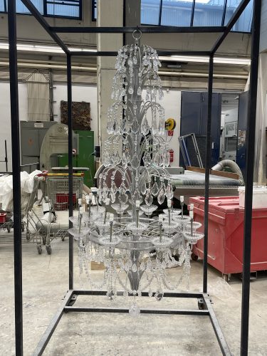 Ein Kronleuchter mit vielen Kristallen hängt in einem Metallgestell in einer Werkstatt oder Fabrikumgebung.