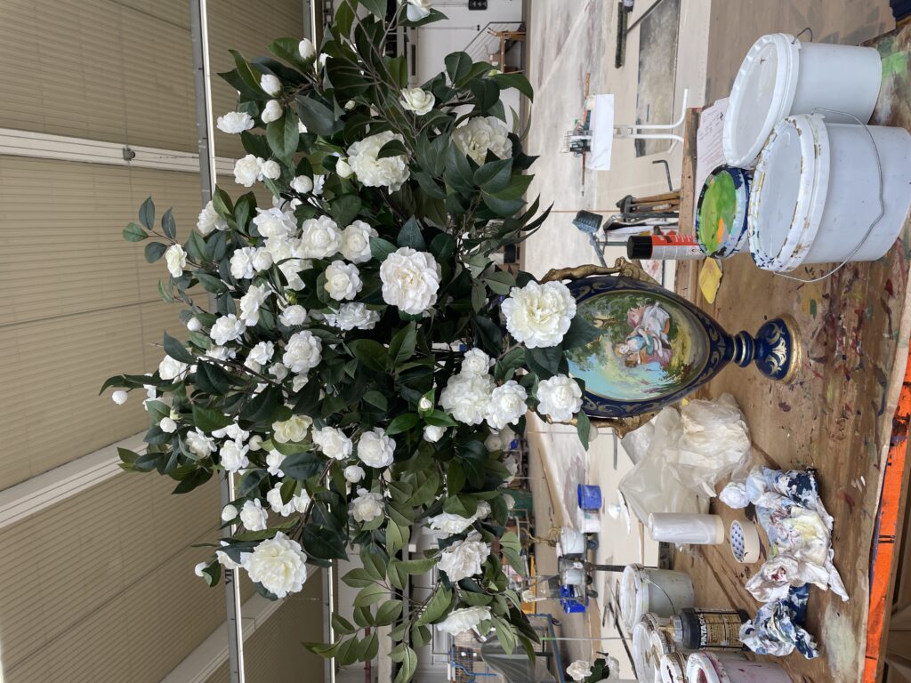 Großer Strauß mit weißen Blumen und grünen Blättern in einer verzierten Vase steht auf einem Werkstatttisch mit Farbdosen und Werkzeug.