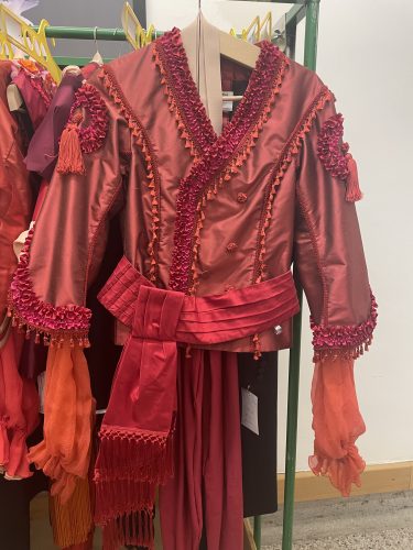 Eine aufwendig verzierte rote Jacke mit Fransendetails und auffälligem Gürtel hängt auf einem Kleiderbügel.
