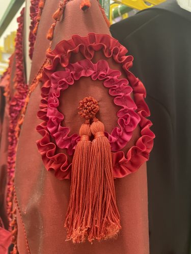 Rote Stoffapplikation mit Rüschen und zwei großen Fransenquasten an einem dunkelroten Kleidungsstück.
