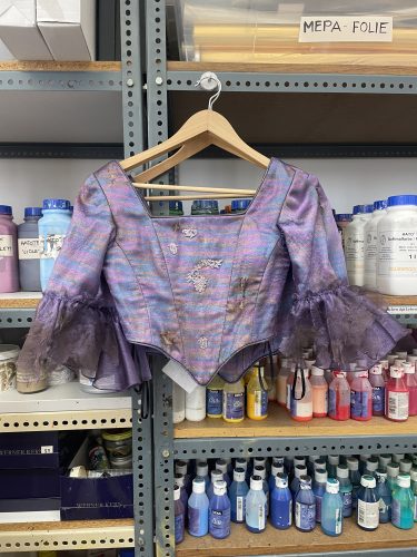 Lila-blaues, glänzendes Oberteil mit weiten transparenten Ärmeln und floralem Muster, hängt auf einem Bügel im Regal mit Farbdosen.