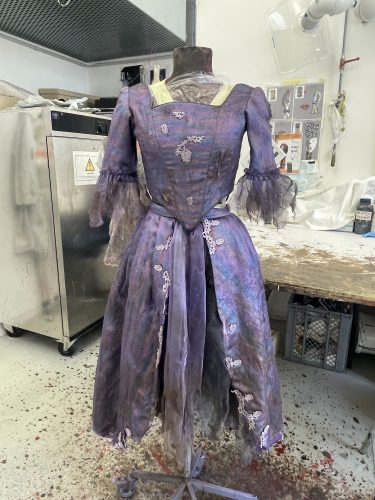 Ein lilafarbenes, viktorianisch inspiriertes Kleid mit Spitzenverzierungen steht auf einer Schneiderpuppe in einer Werkstatt.