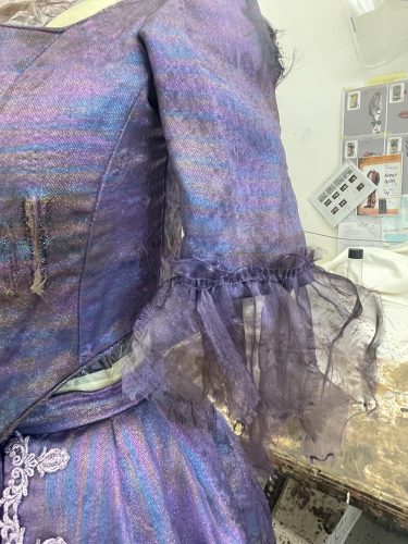 Nahaufnahme eines violett-blauen, schimmernden historischen Kleides mit transparenten, gerüschten Ärmeln und dekorativer Spitze.