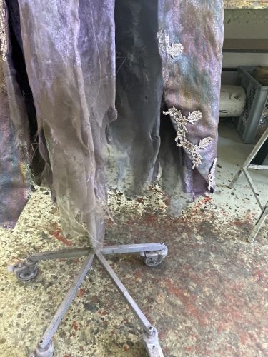 Zerfetztes, staubiges Kleidungsstück an einem Metallständer, verziert mit silbernen Applikationen, auf einem schmutzigen Boden.