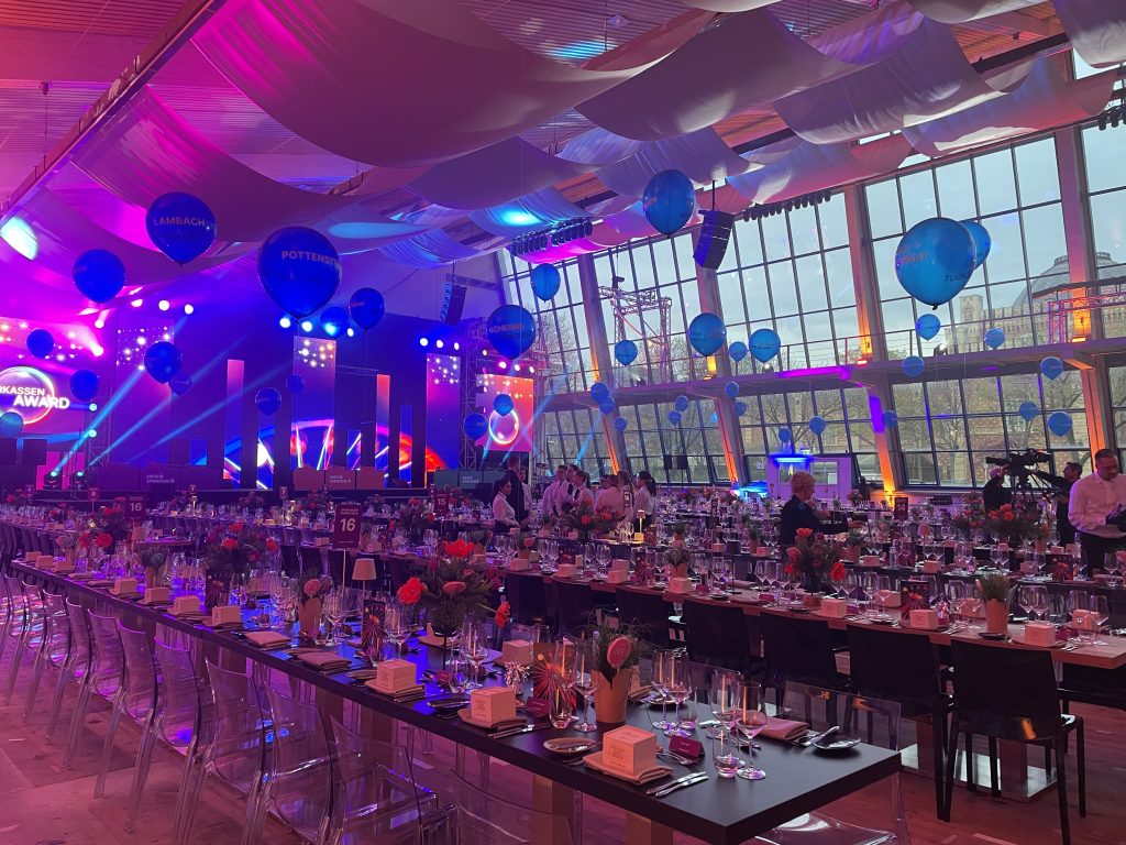 Großer festlich gedeckter Saal mit langen Tischen, Ballons an der Decke und bunter Bühnenbeleuchtung für eine Gala-Veranstaltung.
