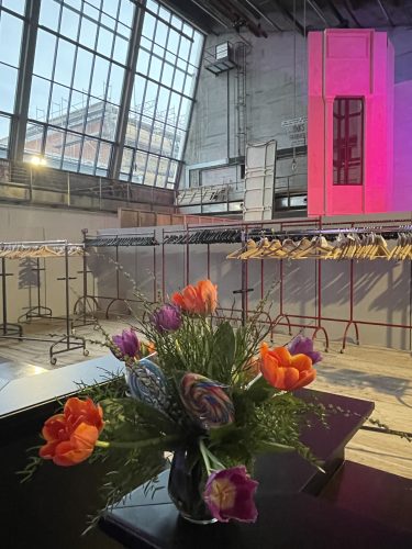 Ein Strauß bunter Tulpen steht auf einem Tisch in einem modernen Raum mit großen Fenstern, Kleiderständern und offenen Garderobenhaken.