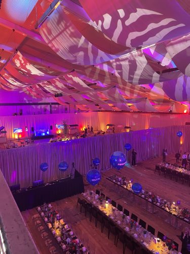 Großer, festlich dekorierter Saal mit langen, gedeckten Tischen, bunten Lichtern und schwebenden blauen Luftballons.