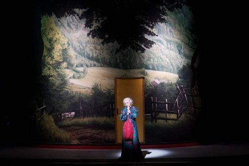 Eine Person in auffälligem Kleid steht allein auf einer Theaterbühne vor einer bemalten ländlichen Kulisse.