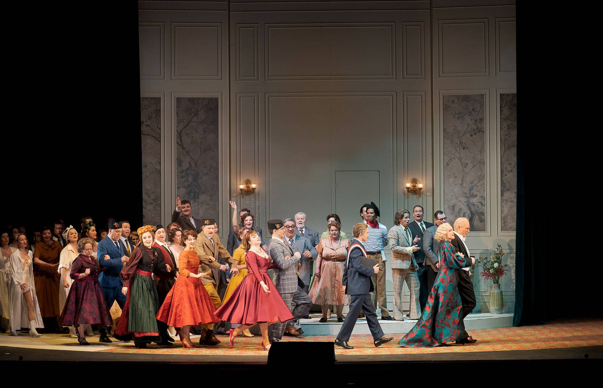 Eine große Gruppe elegant gekleideter Menschen steht und tanzt auf einer Theaterbühne mit festlicher Kulisse.