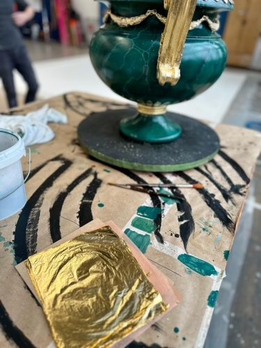 Ein großes grünes Gefäß mit goldenen Verzierungen steht auf einem bemalten Arbeitstisch, daneben liegt ein Blatt Blattgold.