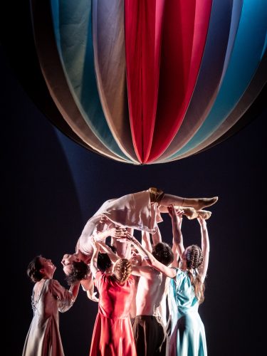 Mehrere Tänzerinnen und Tänzer heben eine Frau unter einem großen, bunten Ballon in einem Theatersaal empor.