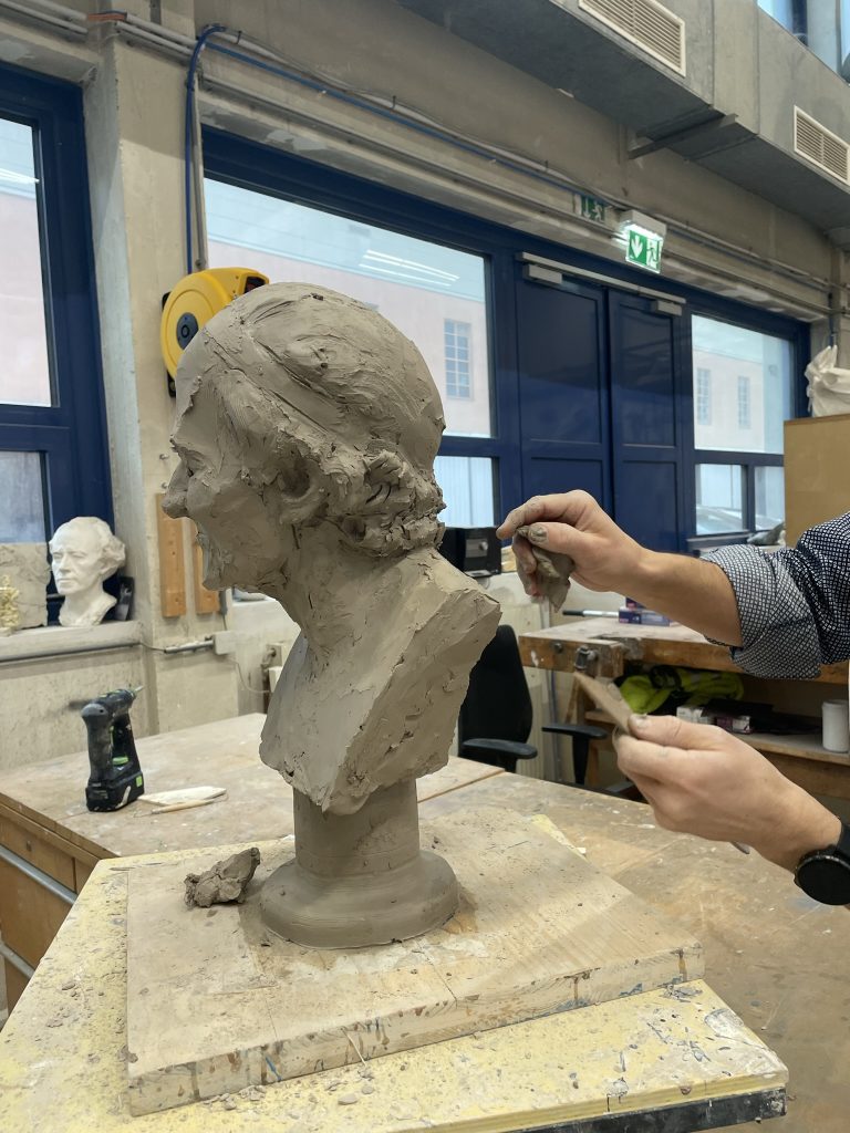 Eine Person modelliert eine Tonbüste einer Frau auf einem Arbeitstisch in einem Atelier.