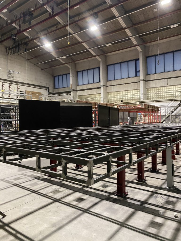 Metallrahmenkonstruktion in einer großen Industriehalle mit hohen Decken und Regalen im Hintergrund.