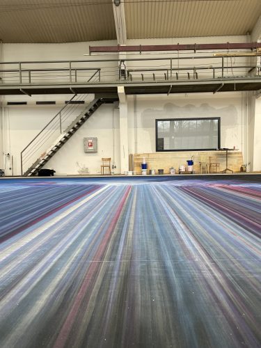 Boden mit bunten, strahlenförmigen Linien in einer Werkhalle, im Hintergrund Treppe, Stühle und Arbeitsmaterialien.