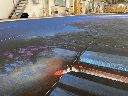 Großflächiges Gemälde mit abstrakten Farbmustern in Blau, Schwarz, Rot und Weiß auf dem Boden eines Ateliers.