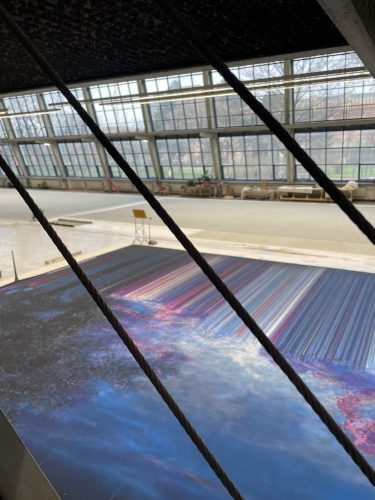 Große Halle mit Panoramafenstern, im Vordergrund ein buntes abstraktes Kunstwerk und eiserne Streben, hinten leere Fläche.