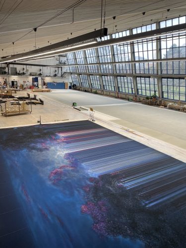 Eine große Halle mit hohen Fenstern, im Vordergrund ein digital bedruckter Stoff mit abstraktem, blau-violettem Muster.