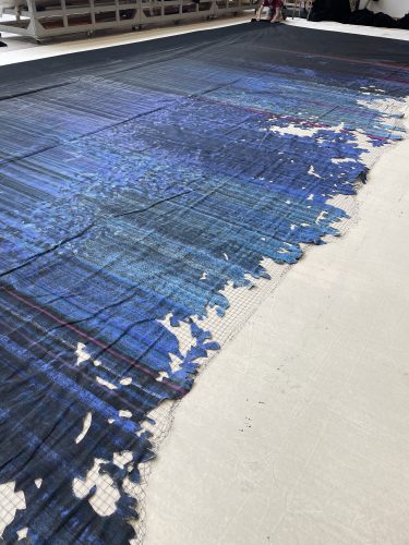 Ein großes textilähnliches Gewebe mit blauem und schwarzem Muster liegt ausgebreitet auf dem Boden, am Rand ist es stark ausgefranst.