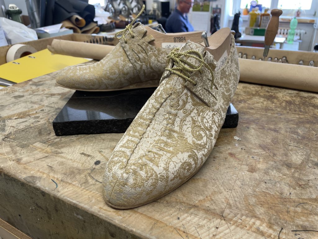 Ein Paar elegante, gemusterte Schnürschuhe mit gold-weißen Verzierungen steht auf einer Werkbank in einer Werkstatt.