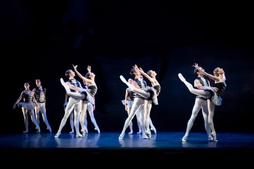 Mehrere Balletttänzerinnen und -tänzer führen auf einer Bühne synchron elegante Bewegungen in schwarzen und weißen Kostümen aus.