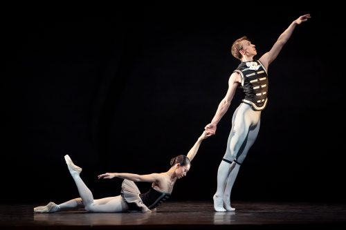 Zwei Balletttänzer auf der Bühne, ein Mann steht mit erhobenem Arm, während eine Frau elegant auf dem Boden liegt.