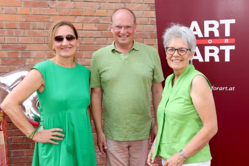 Zwei Frauen und ein Mann lächeln vor einer Backsteinwand und einem Banner mit der Aufschrift „ART FOR ART“.