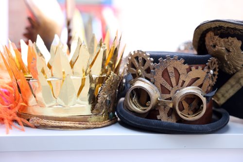 Goldene Krone und schwarzer Steampunk-Hut mit Zahnrädern und Schutzbrille liegen nebeneinander auf einer weißen Oberfläche.