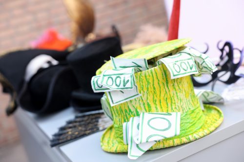 Ein auffälliger gelb-grüner Zylinder mit Spielgeldscheinen darauf liegt auf einem Tisch, im Hintergrund weitere bunte Hüte.