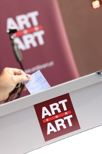 Eine Hand hält einen Zettel über eine Box mit dem Aufdruck „ART FOR ART“, im Hintergrund ein unscharfes gleiches Logo.