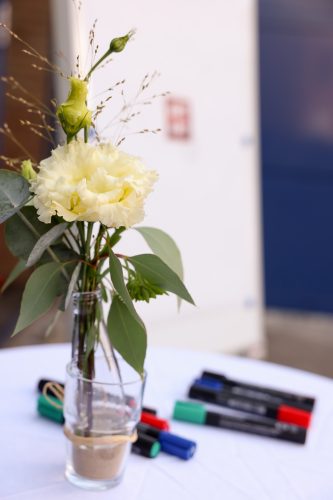 Eine weiße Blume in einer Glasvase mit Sand steht auf einem runden Tisch, im Hintergrund liegen bunte Filzstifte.