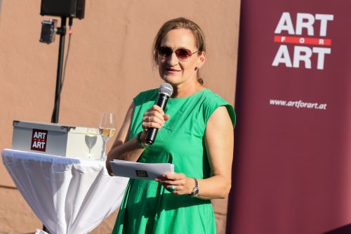 Frau in grünem Kleid spricht mit Mikrofon und hält einen Zettel, im Hintergrund Banner und Tisch mit Weinglas und „ART FOR ART“-Logo.