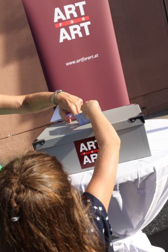 Ein Kind wirft eine Karte in eine silberne Box mit der Aufschrift "ART FOR ART" vor einem passenden Banner ein.