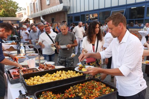 Menschen bedienen sich an einem Buffet im Freien mit verschiedenen warmen Speisen wie Kartoffeln und Gemüse.