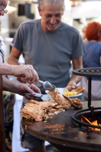 Zwei Personen grillen Fleisch auf einer Feuerplatte, eine hält Zange und Gabel, im Hintergrund lächelt ein Mann.