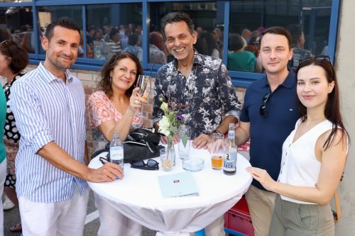 Fünf Menschen stehen lächelnd um einen Stehtisch mit Getränken und Blumen bei einer Veranstaltung im Freien.
