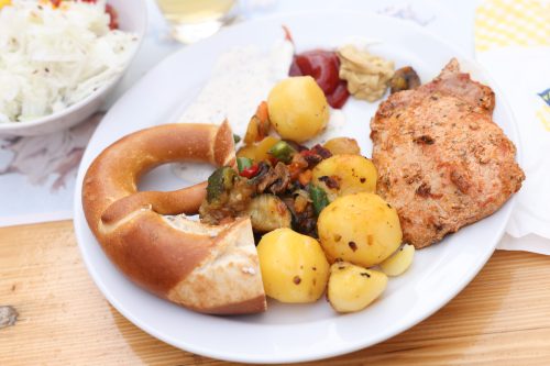 Ein Teller mit Schweinesteak, Pellkartoffeln, gebratenem Gemüse und einer Brezel, dazu verschiedene Soßen.