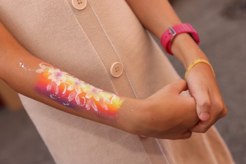 Ein Kind mit bemaltem Unterarm, darauf bunte Blumen und Blätter, trägt ein beiges Kleid und ein pinkes Armband.
